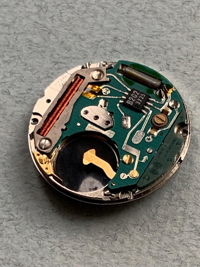 May include: Close-up of a watch movement with a green circuit board, copper coil, and a small black component labeled "B202 3235". The watch mechanism is circular and has various metal components.