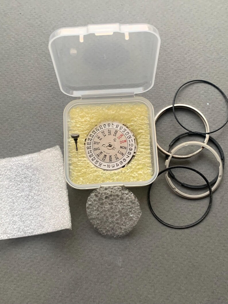 Pu&ograve; includere: Kit di riparazione orologi con vari componenti. Include un movimento dell'orologio con visualizzazione del giorno e della data, una custodia di plastica trasparente, una piccola vite, un filtro in schiuma e diverse guarnizioni per orologi nere e argentate.