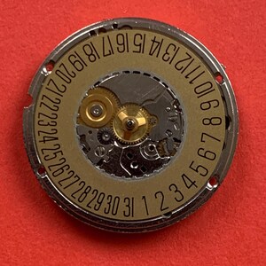 May include: A close-up of a watch movement with a gold-colored dial and silver-colored metal casing. The dial features the numbers 1 through 31, indicating the days of the month. The intricate gears and components are visible.