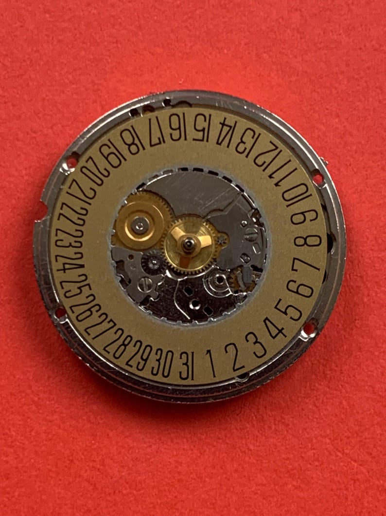 May include: A close-up of a watch movement with a gold-colored dial and silver-colored metal casing. The dial features the numbers 1 through 31, indicating the days of the month. The intricate gears and components are visible.