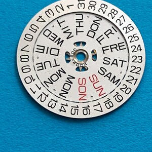 May include: A circular watch calendar dial with a white face and black text. The days of the week are printed around the center, with "SUN" in red. Numbers 1-31 are around the outer edge. The dial has a central hole.
