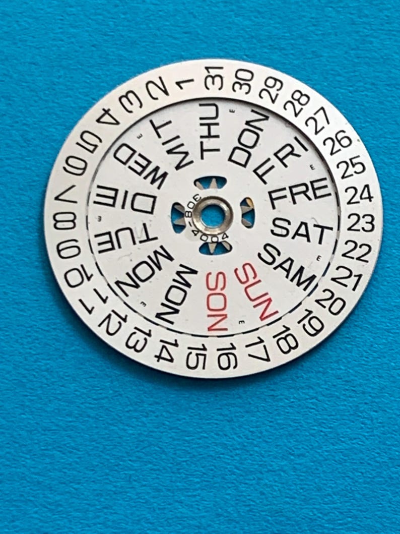 May include: A circular watch calendar dial with a white face and black text. The days of the week are printed around the center, with "SUN" in red. Numbers 1-31 are around the outer edge. The dial has a central hole.