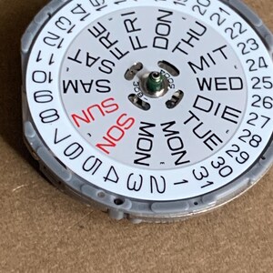 May include: A watch calendar dial with a white face, featuring the days of the week and numbers 1-31. The days of the week are in black, except for Sunday, which is in red. The dial is encased in a gray metal frame.
