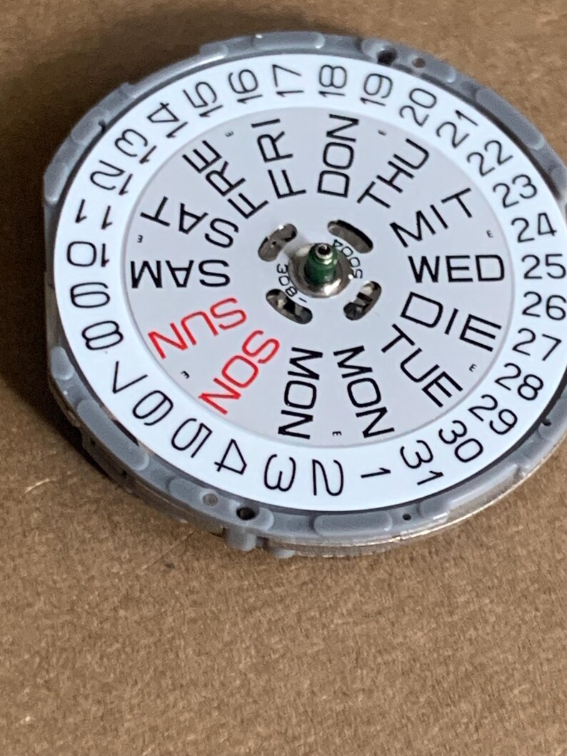 May include: A watch calendar dial with a white face, featuring the days of the week and numbers 1-31. The days of the week are in black, except for Sunday, which is in red. The dial is encased in a gray metal frame.