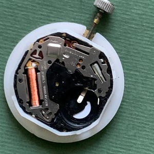 K&ouml;nnte beinhalten: Ein Uhrwerk mit wei&szlig;em Kunststoffgeh&auml;use, das komplizierte Metallkomponenten, eine kupferfarbene Spule und eine silberne Krone aufweist. Der Uhrenmechanismus ist f&uuml;r Reparatur oder Ersatzteile konzipiert.