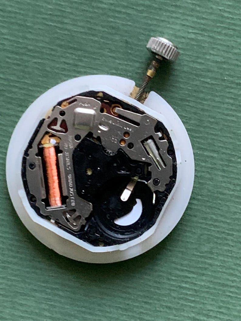 K&ouml;nnte beinhalten: Ein Uhrwerk mit wei&szlig;em Kunststoffgeh&auml;use, das komplizierte Metallkomponenten, eine kupferfarbene Spule und eine silberne Krone aufweist. Der Uhrenmechanismus ist f&uuml;r Reparatur oder Ersatzteile konzipiert.