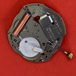K&ouml;nnte beinhalten: Nahaufnahme eines Uhrwerks mit der Aufschrift "15700 JAPAN" und "ORIENT WATCH CO. LTD.". Das Uhrwerk hat eine Kupferspule, eine wei&szlig;e runde Komponente und eine goldfarbene Schraube. Der Hintergrund ist rot.