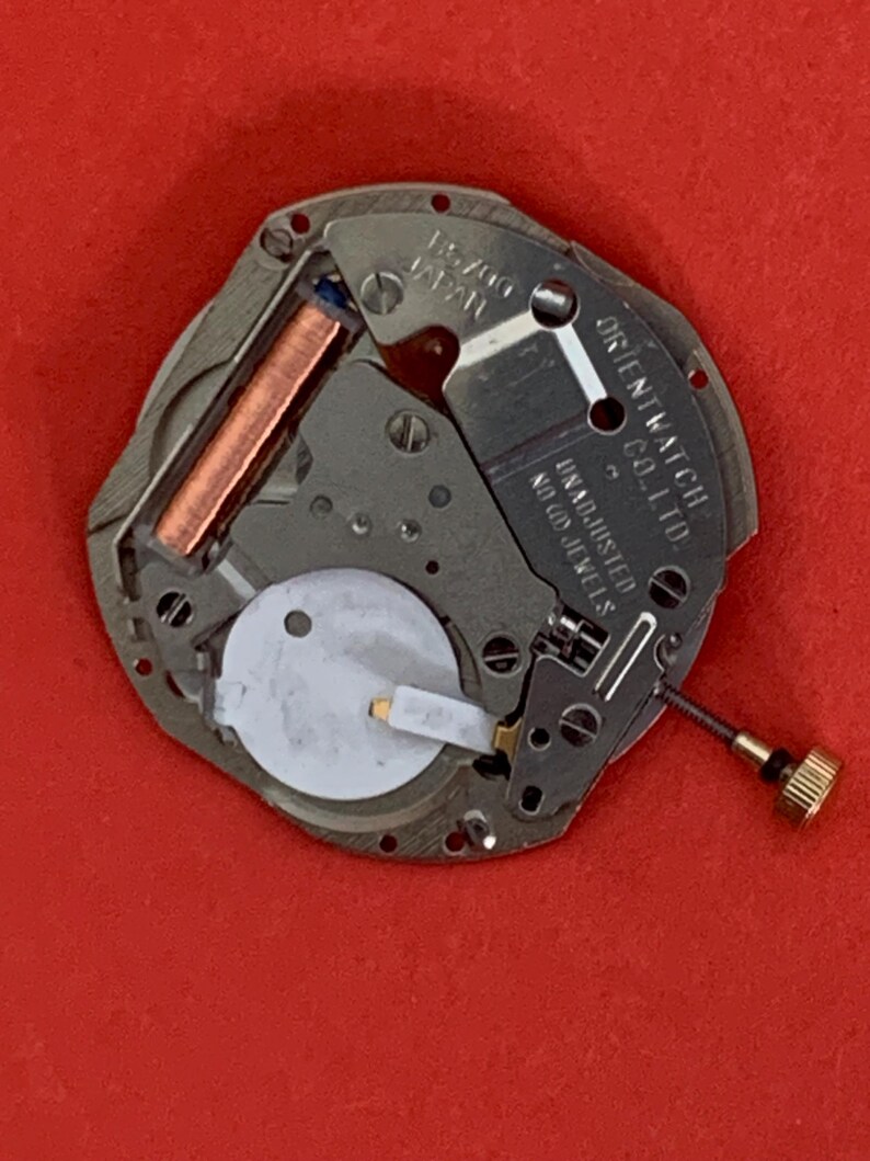 K&ouml;nnte beinhalten: Nahaufnahme eines Uhrwerks mit der Aufschrift "15700 JAPAN" und "ORIENT WATCH CO. LTD.". Das Uhrwerk hat eine Kupferspule, eine wei&szlig;e runde Komponente und eine goldfarbene Schraube. Der Hintergrund ist rot.