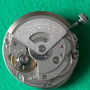 May include: Close-up of a watch movement with intricate gears and components. The silver-toned mechanism features the text "OPIENT WATCH CO., LTD. JAPAN". A small, silver crown is visible, along with jewels and screws. The watch movement is on a green surface.