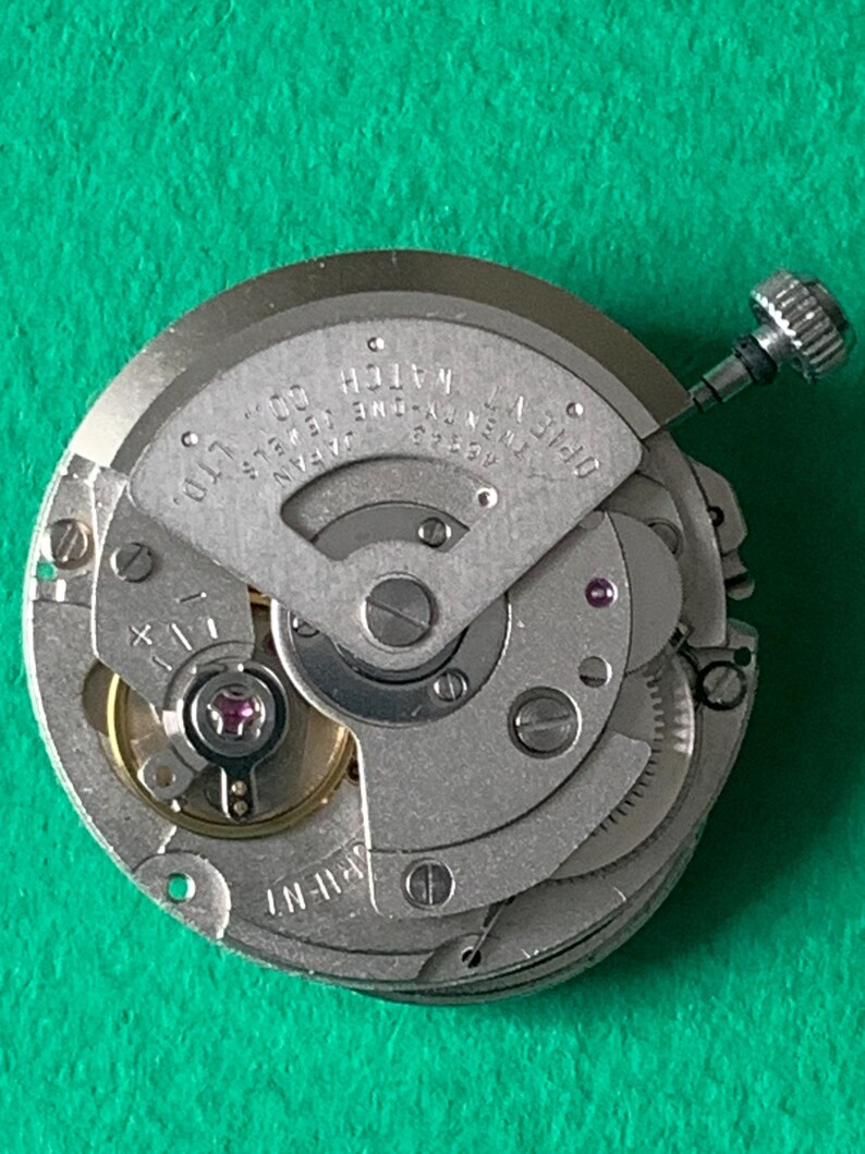 May include: Close-up of a watch movement with intricate gears and components. The silver-toned mechanism features the text "OPIENT WATCH CO., LTD. JAPAN". A small, silver crown is visible, along with jewels and screws. The watch movement is on a green surface.