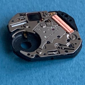 May include: Close-up of a watch movement, showcasing intricate mechanical components. The movement features silver-colored metal parts, a black circular component, and a copper-colored coil. The background is a solid light blue.
