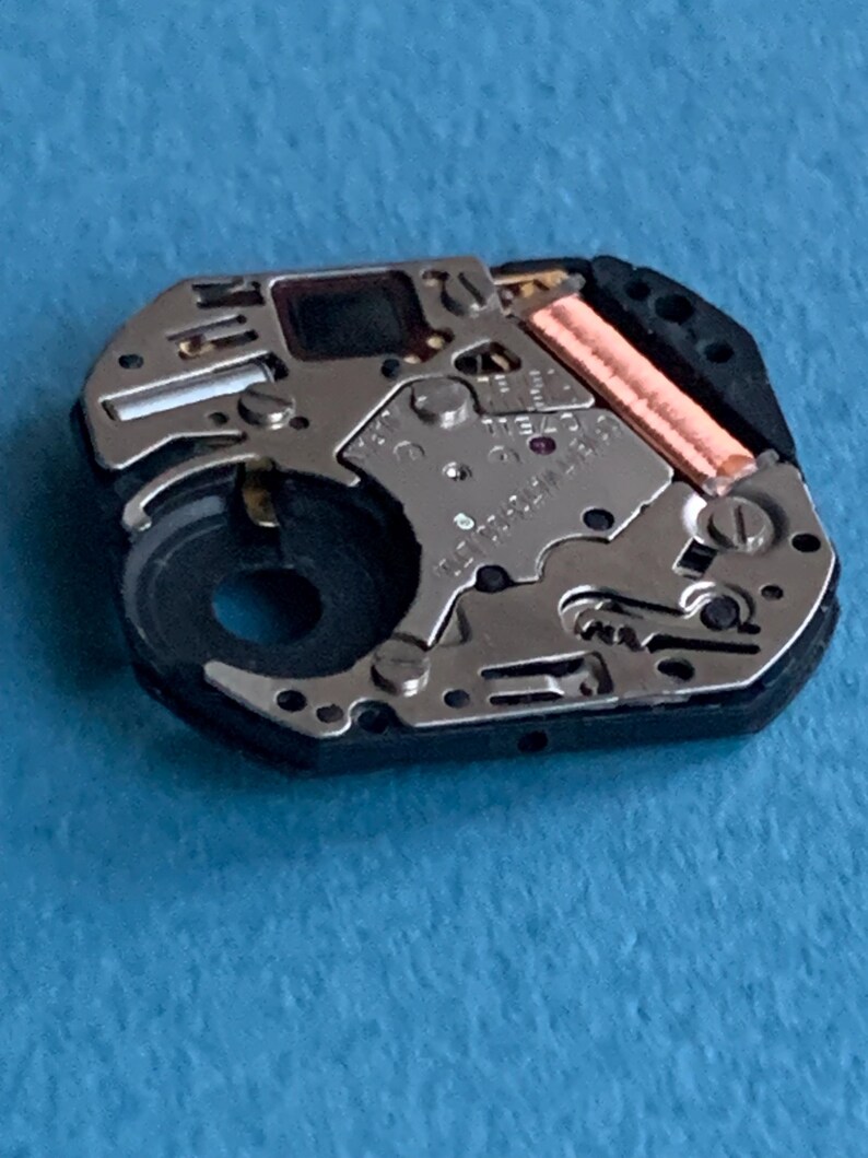 May include: Close-up of a watch movement, showcasing intricate mechanical components. The movement features silver-colored metal parts, a black circular component, and a copper-colored coil. The background is a solid light blue.