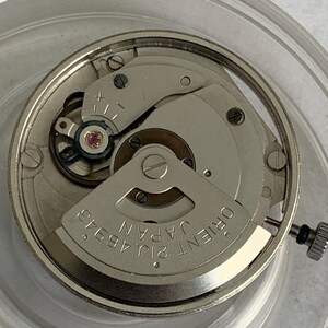 May include: Close-up of a silver-toned watch movement with intricate gears and components. The watch mechanism features the text "ORIENT JAPAN 21J 46943". The image is taken from above, showcasing the detailed inner workings of the watch.