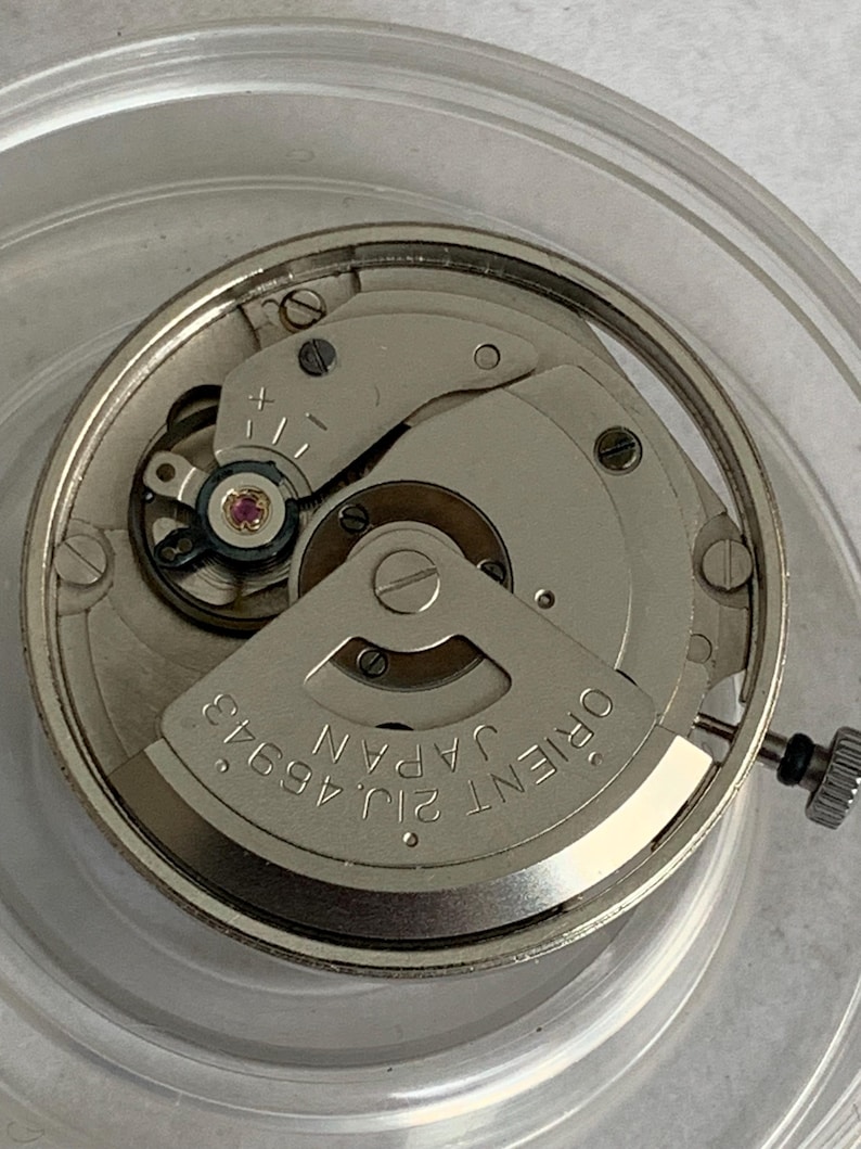 May include: Close-up of a silver-toned watch movement with intricate gears and components. The watch mechanism features the text "ORIENT JAPAN 21J 46943". The image is taken from above, showcasing the detailed inner workings of the watch.