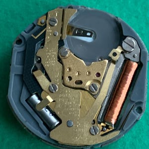 May include: Close-up of a watch movement with gold-colored gears, a copper coil, and a silver battery. The movement is set in a gray plastic casing. The background is a solid green color.