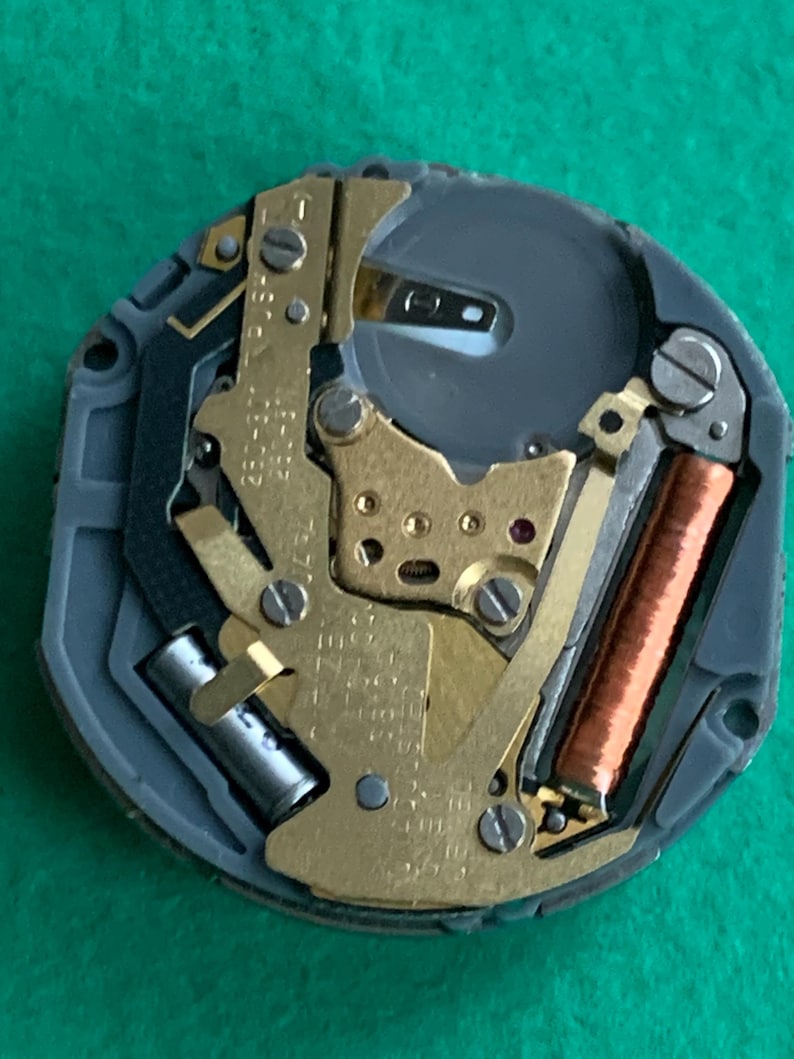 May include: Close-up of a watch movement with gold-colored gears, a copper coil, and a silver battery. The movement is set in a gray plastic casing. The background is a solid green color.