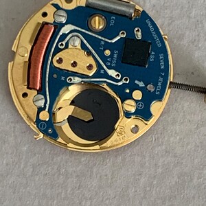 May include: A close-up of a watch movement with a gold-colored frame and a blue circuit board. The board has silver lines, a black battery, and the text "UNADJUSTED" and "SWISS". The movement has seven jewels.