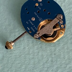 May include: A close-up of a watch movement with a blue circuit board. The movement has gold-colored gears and a silver lever. The text "SWISS SIX 6 JEWELS" and "V8" are visible. A gold-colored crown is attached to a threaded rod.