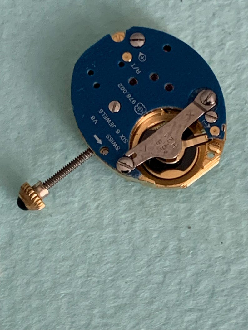 May include: A close-up of a watch movement with a blue circuit board. The movement has gold-colored gears and a silver lever. The text "SWISS SIX 6 JEWELS" and "V8" are visible. A gold-colored crown is attached to a threaded rod.