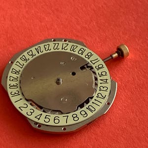 K&ouml;nnte beinhalten: Eine Uhrwerkkomponente mit einer silberfarbenen Metallbasis und einem wei&szlig;en Ring, der die Tage des Monats von 1 bis 31 anzeigt. Eine kleine goldfarbene Schraube ist seitlich angebracht. Der Hintergrund ist rot.
