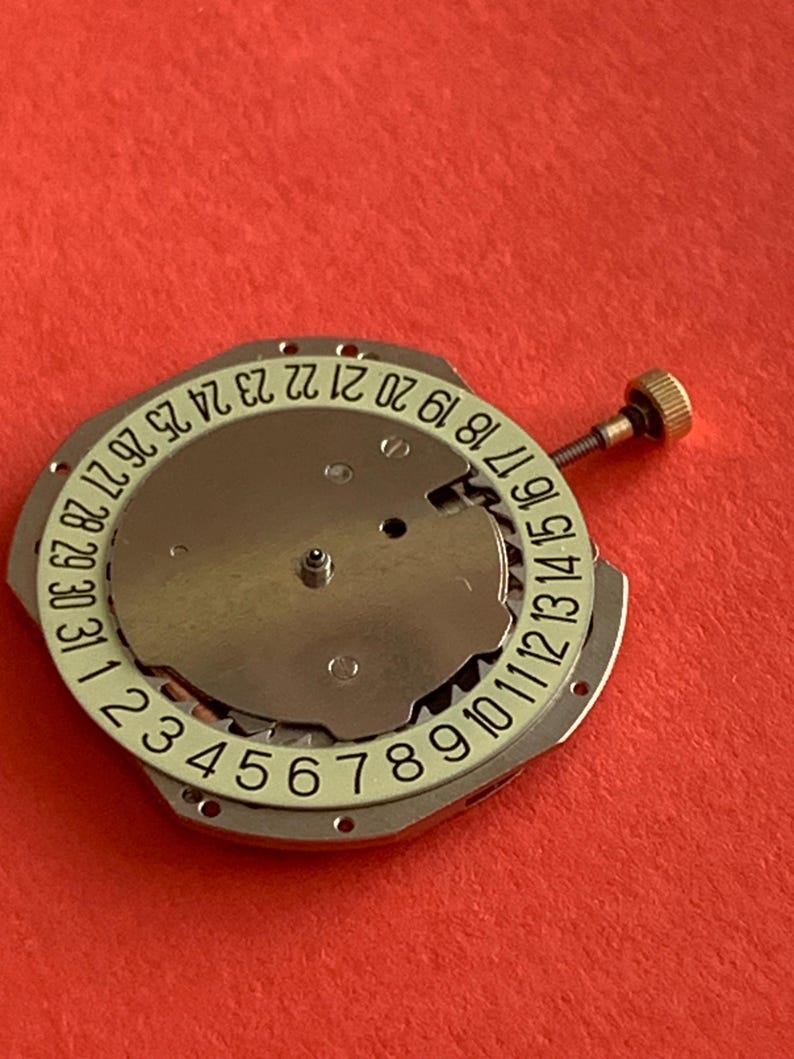 K&ouml;nnte beinhalten: Eine Uhrwerkkomponente mit einer silberfarbenen Metallbasis und einem wei&szlig;en Ring, der die Tage des Monats von 1 bis 31 anzeigt. Eine kleine goldfarbene Schraube ist seitlich angebracht. Der Hintergrund ist rot.