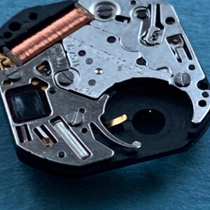 May include: Close-up of a watch movement, showcasing intricate mechanical components. The image highlights the metallic gears, screws, and a copper coil. The watch mechanism is set against a blue background, emphasizing the precision of the watchmaking.