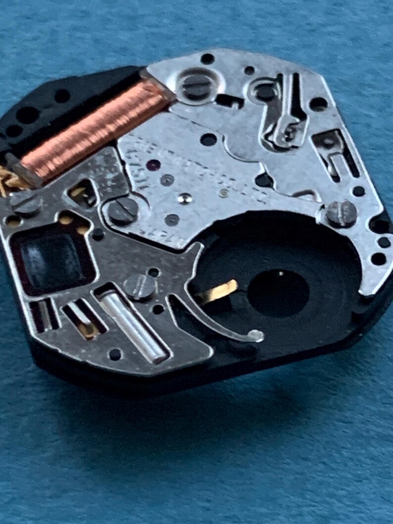 May include: Close-up of a watch movement, showcasing intricate mechanical components. The image highlights the metallic gears, screws, and a copper coil. The watch mechanism is set against a blue background, emphasizing the precision of the watchmaking.