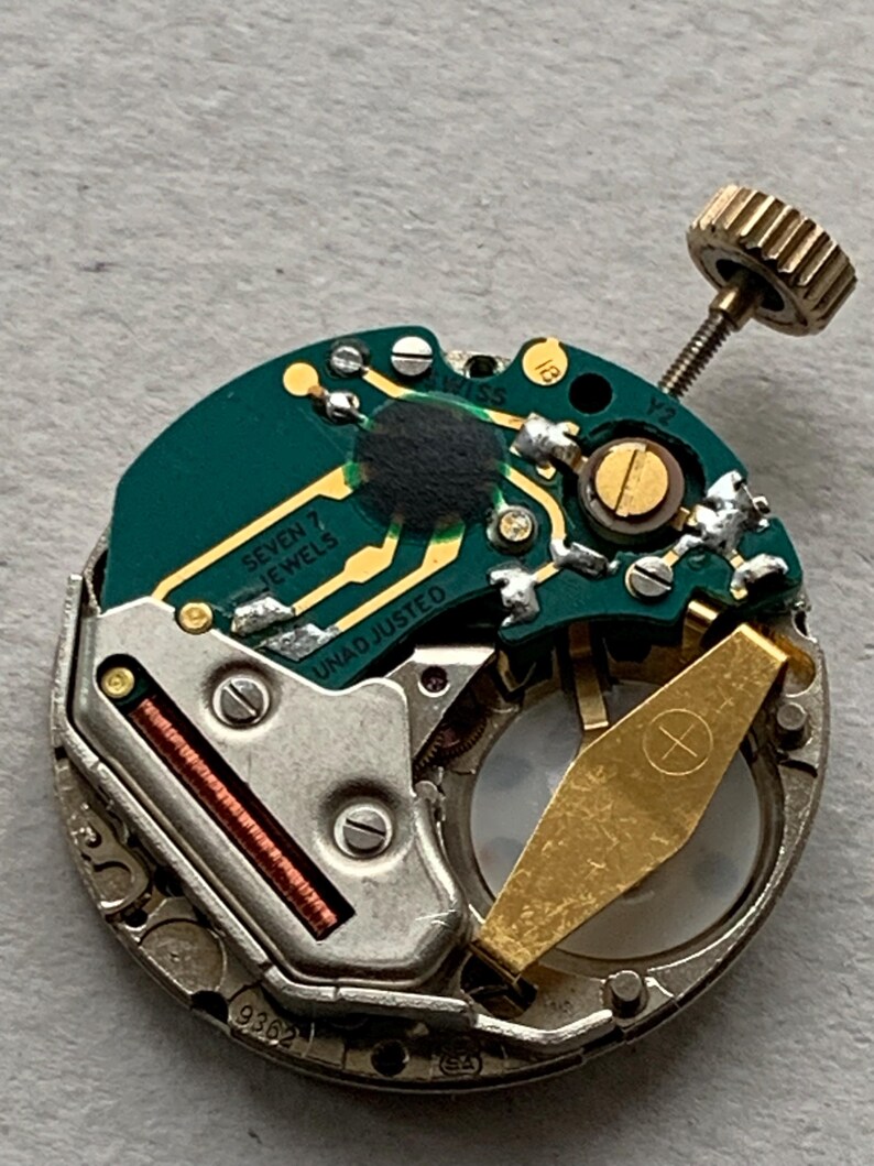 Puede incluir: Primer plano de un movimiento de reloj con una placa de circuito verde. La placa tiene componentes dorados, incluyendo un soporte de bater&iacute;a y una corona. El texto "SEVEN JEWELS" y "UNADJUSTED" son visibles. Componente mec&aacute;nico de reloj.