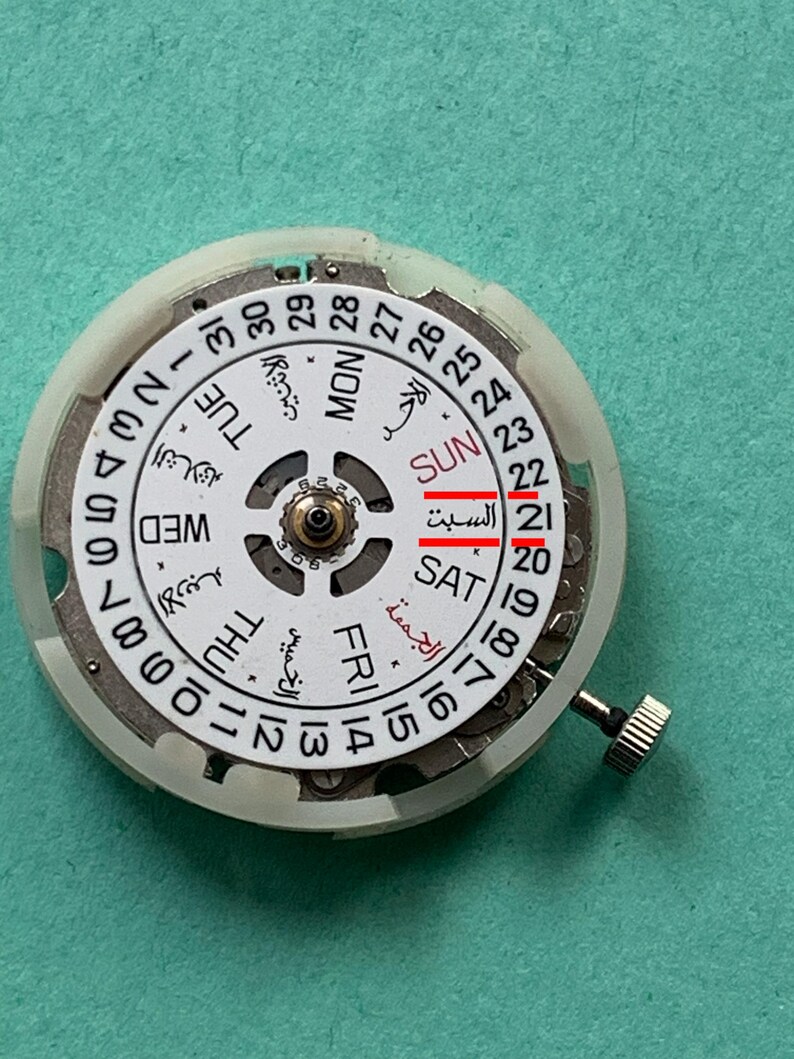 May include: A close-up of a watch movement with a white dial and a silver-colored metal casing. The dial displays the days of the week in English and Arabic, with the date highlighted. The watch crown is visible on the right side.