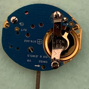May include: A close-up of a watch movement with a blue circuit board and gold-colored components. The watch movement has the text "SWISS", "V8", and "SIX 6 JEWELS". The movement also has a small screw and a metal arm.