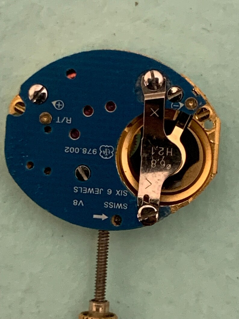 May include: A close-up of a watch movement with a blue circuit board and gold-colored components. The watch movement has the text "SWISS", "V8", and "SIX 6 JEWELS". The movement also has a small screw and a metal arm.