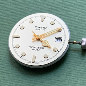 K&ouml;nnte beinhalten: Ein Casio Quartz Zifferblatt mit cremefarbenem Zifferblatt, goldfarbenen Zeigern und Markierungen sowie einem Datumsfenster mit der Zahl 26. Das Zifferblatt tr&auml;gt die Aufschrift "CASIO QUARTZ" und "WATER RESIST 50 M".