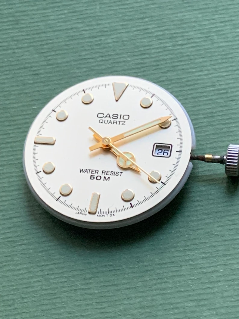 K&ouml;nnte beinhalten: Ein Casio Quartz Zifferblatt mit cremefarbenem Zifferblatt, goldfarbenen Zeigern und Markierungen sowie einem Datumsfenster mit der Zahl 26. Das Zifferblatt tr&auml;gt die Aufschrift "CASIO QUARTZ" und "WATER RESIST 50 M".
