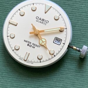 K&ouml;nnte beinhalten: Ein Casio Quartz Zifferblatt mit cremefarbenem Zifferblatt, goldfarbenen Zeigern und Stundenmarkierungen. Das Zifferblatt zeigt "CASIO QUARTZ" und "WATER RESIST 50 M". Ein Datumsfenster zeigt die Zahl 26. Die Uhr hat eine silberfarbene Krone.