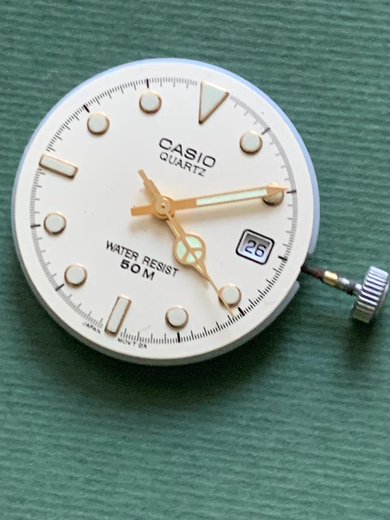 K&ouml;nnte beinhalten: Ein Casio Quartz Zifferblatt mit cremefarbenem Zifferblatt, goldfarbenen Zeigern und Stundenmarkierungen. Das Zifferblatt zeigt "CASIO QUARTZ" und "WATER RESIST 50 M". Ein Datumsfenster zeigt die Zahl 26. Die Uhr hat eine silberfarbene Krone.