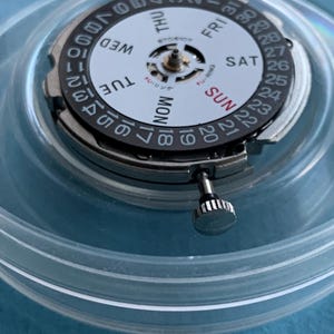 May include: A detailed view of a watch movement, featuring a white dial with black numerals and the days of the week. The movement is housed in a clear plastic container. The crown is silver.