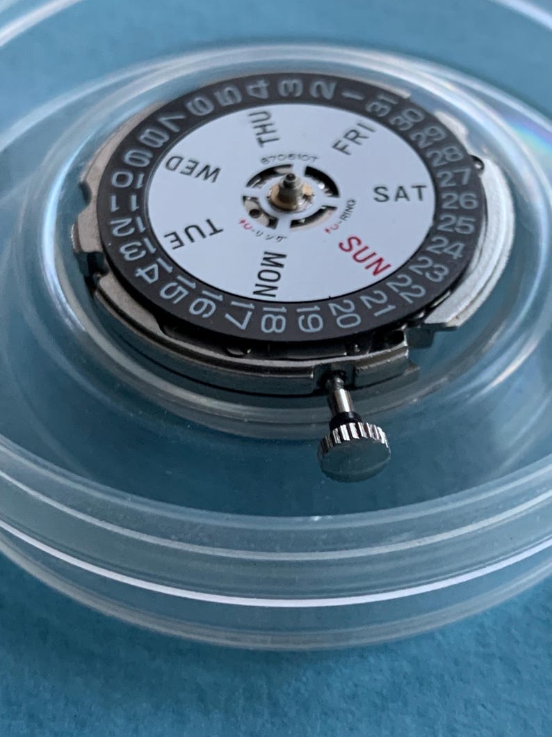 May include: A detailed view of a watch movement, featuring a white dial with black numerals and the days of the week. The movement is housed in a clear plastic container. The crown is silver.
