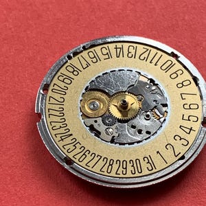 May include: Close-up of a watch movement with a gold-colored date wheel and silver-colored gears. The date wheel has numbers from 1 to 31. The background is a solid red color.