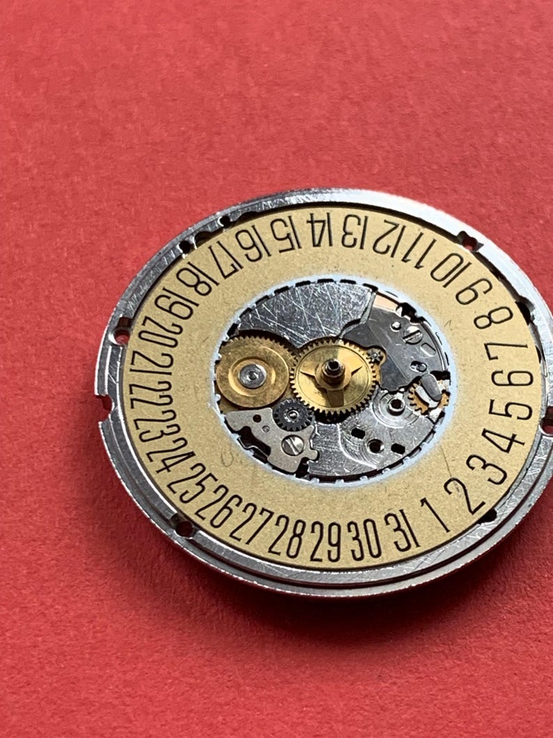 May include: Close-up of a watch movement with a gold-colored date wheel and silver-colored gears. The date wheel has numbers from 1 to 31. The background is a solid red color.