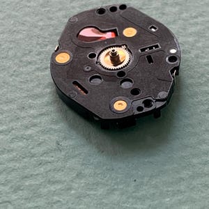 May include: A black watch movement featuring a gold gear and gold contact points. The mechanism includes a red component, set against a light green backdrop. This is a close-up view of the watch's internal workings, showcasing its intricate design.
