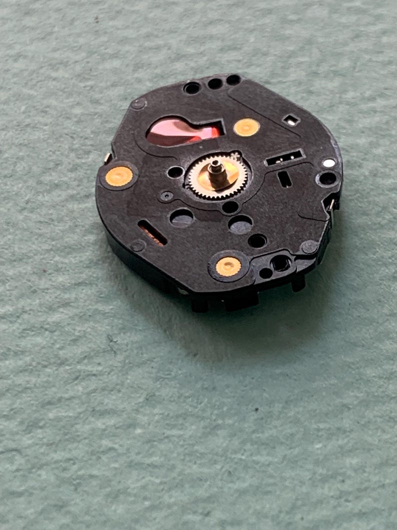 May include: A black watch movement featuring a gold gear and gold contact points. The mechanism includes a red component, set against a light green backdrop. This is a close-up view of the watch's internal workings, showcasing its intricate design.