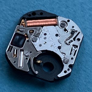 May include: Close-up of a watch movement with intricate metal components, including a copper coil and various gears. The watch movement is black and silver, with the text "ORIENT WATCH CO. LTD." visible.