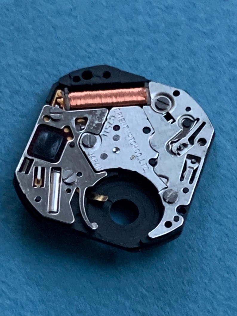 May include: Close-up of a watch movement with intricate metal components, including a copper coil and various gears. The watch movement is black and silver, with the text "ORIENT WATCH CO. LTD." visible.