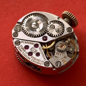 May include: Close-up of a vintage Longines watch movement with intricate gears, jewels, and the text "Longines Watch Co. Swiss Seventeen 17 Jewels". The watch mechanism is set against a red background.