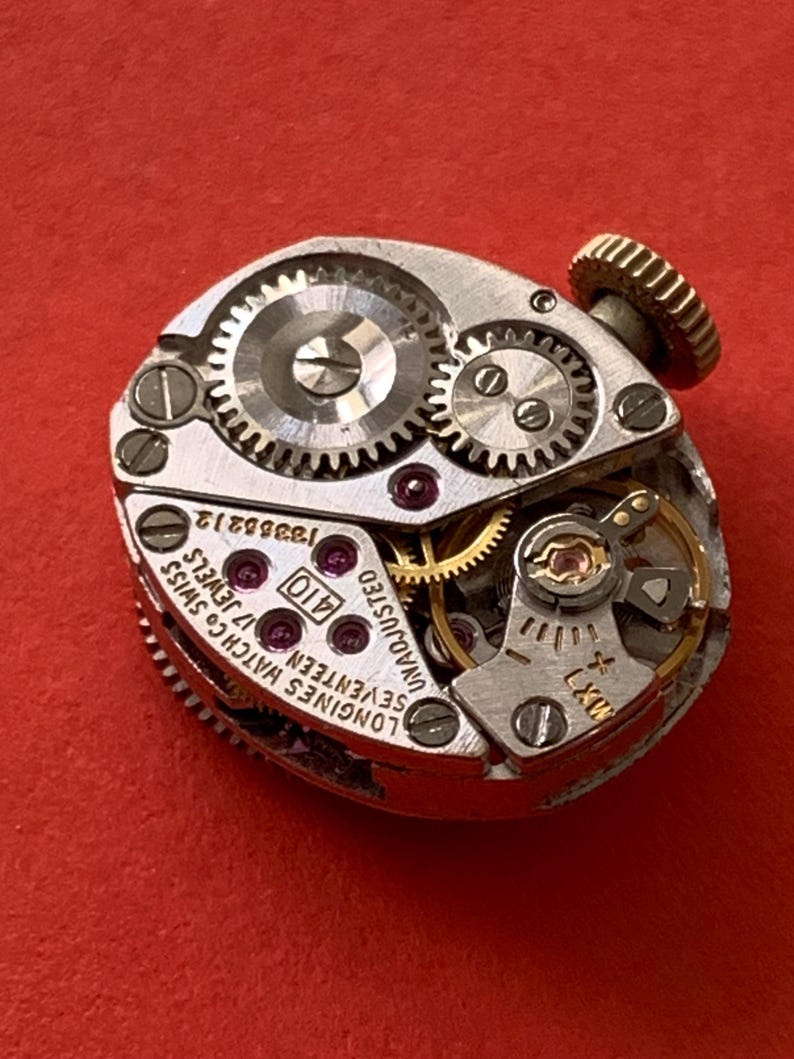May include: Close-up of a vintage Longines watch movement with intricate gears, jewels, and the text "Longines Watch Co. Swiss Seventeen 17 Jewels". The watch mechanism is set against a red background.