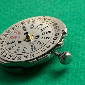 May include: Close-up of a silver watch movement with a day and date dial. The dial displays the days of the week and numbers for the date. A small crown is visible on the side. The background is a solid green.