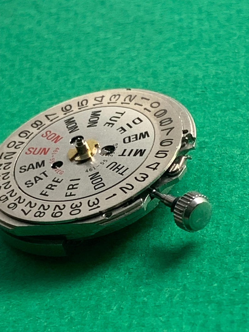 May include: Close-up of a silver watch movement with a day and date dial. The dial displays the days of the week and numbers for the date. A small crown is visible on the side. The background is a solid green.