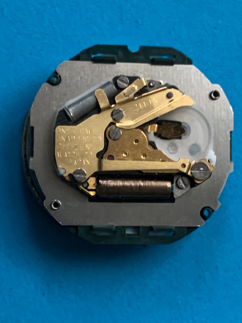 May include: A watch movement featuring gold-coloured gears and components, including a coil and a balance wheel. The movement is mounted on a silver-coloured base with a green plastic frame. The text "ONE JEWEL UNADJUSTED CITIZEN JAPAN" is visible.