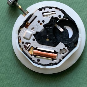 K&ouml;nnte beinhalten: Nahaufnahme eines Uhrwerks mit wei&szlig;em Kunststoffgeh&auml;use. Die inneren Komponenten umfassen silberne Metallteile, eine kupferfarbene Spule und eine silberne Krone. Der Hintergrund ist einfarbig gr&uuml;n.