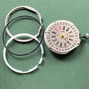 May include: Watch movement components, including a dial with day and date displays, and three circular rings. The dial shows days of the week in English and German, and numbers 1-31. The parts are on a green surface.